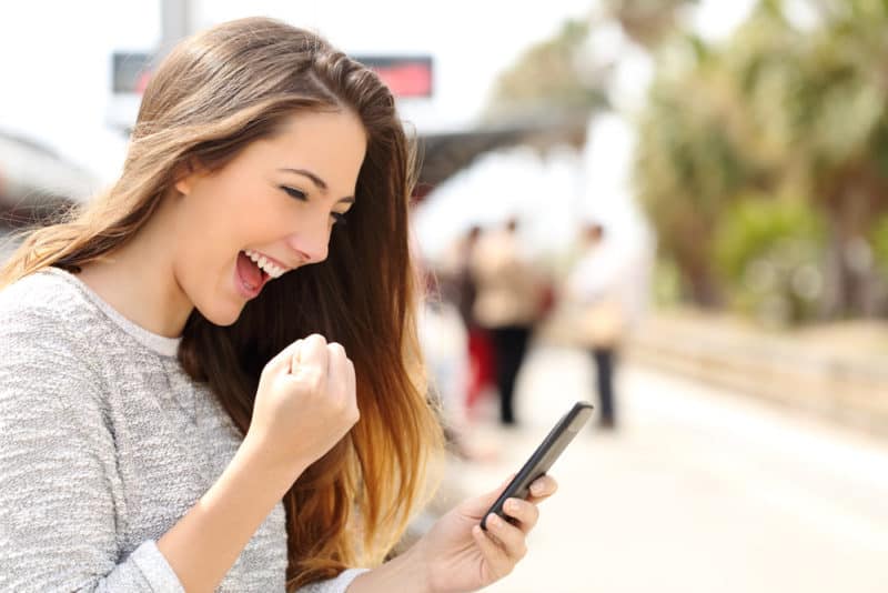 Los mejores sitios de encuestas mujer joven afuera con el teléfono en la mano felizmente haciendo un puño y sonriendo al teléfono